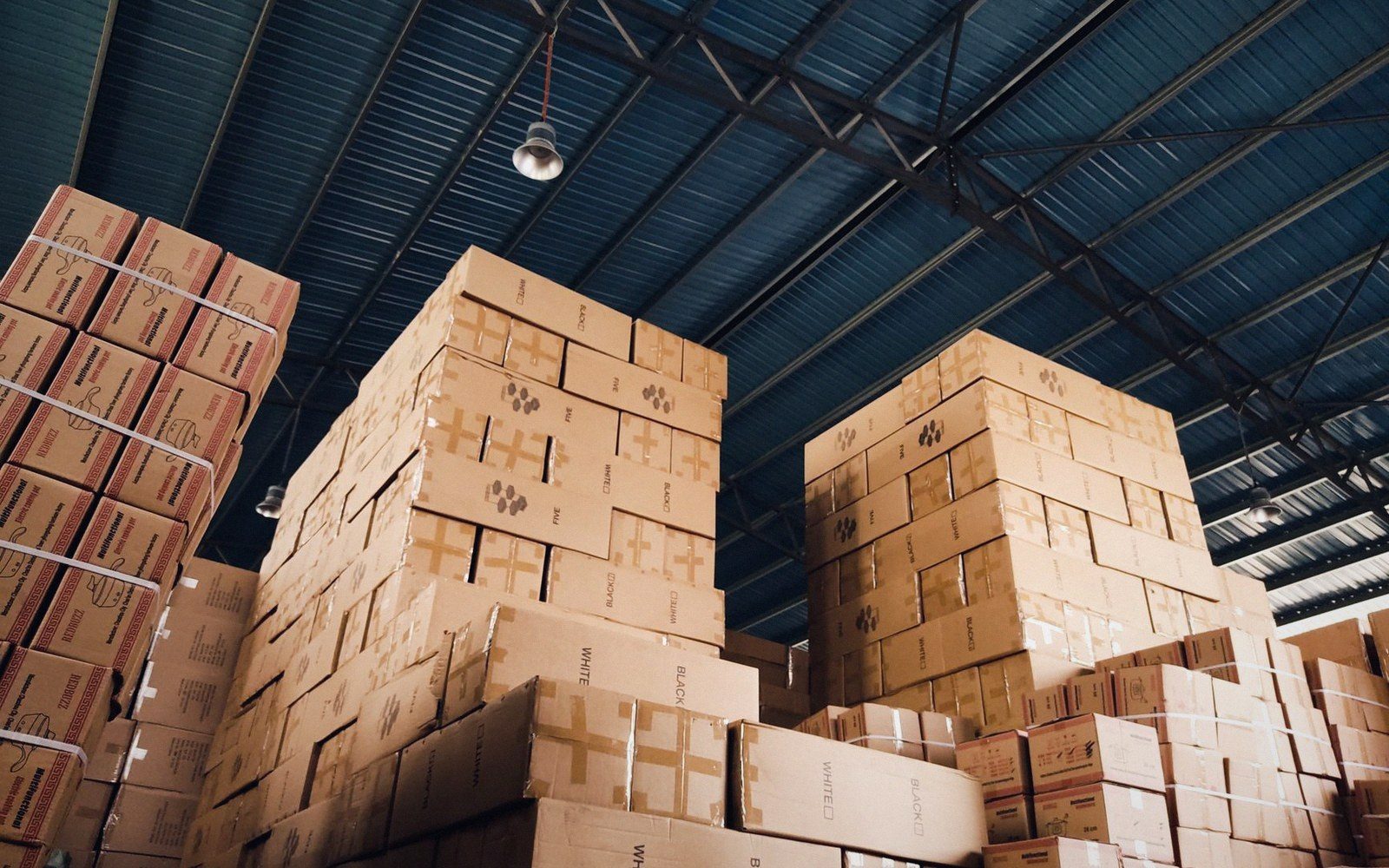 Stacks of cardboard boxes towering under a metal-roofed warehouse ceiling