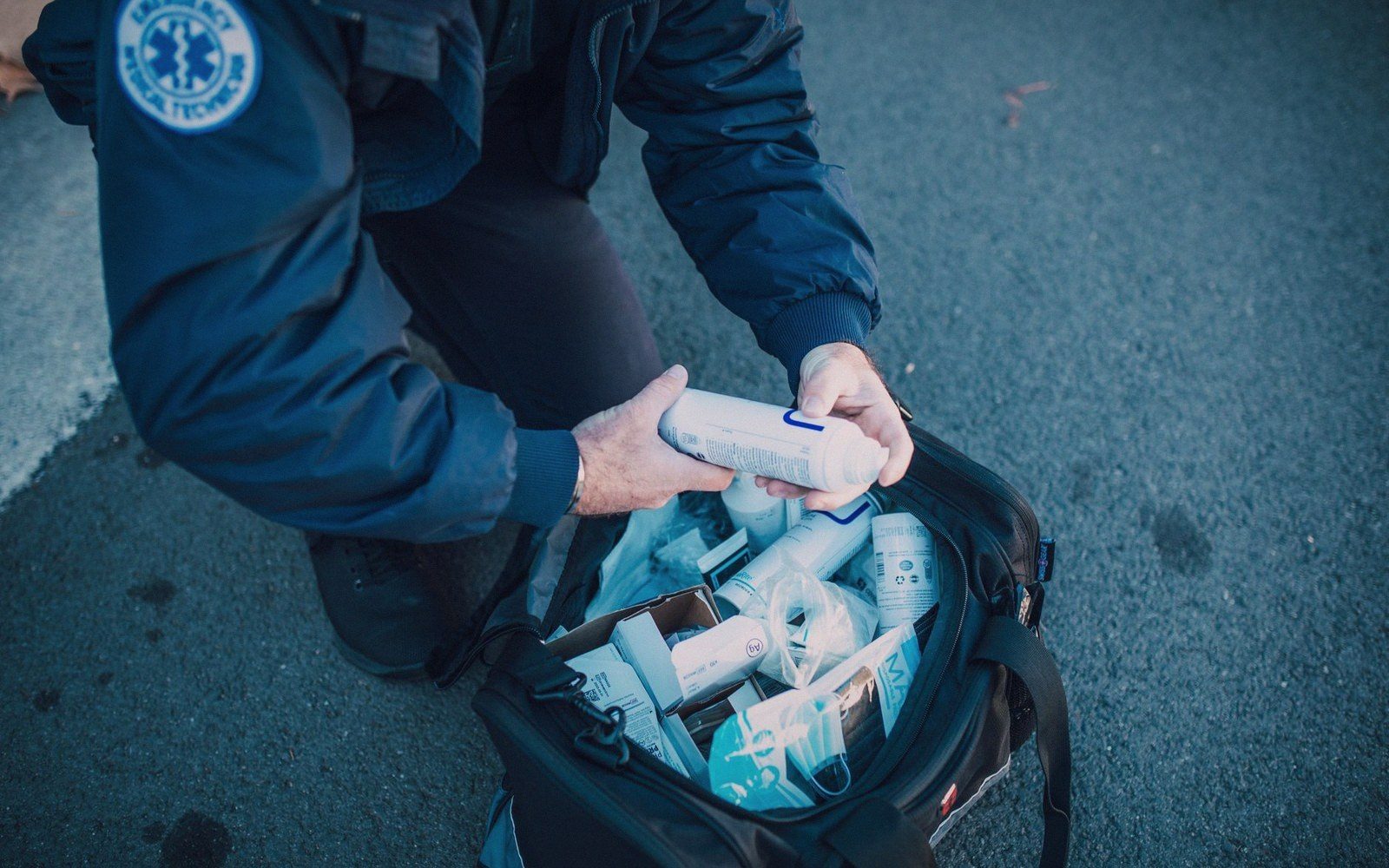 An EMT kneels on pavement, packing medical supplies into an open emergency response bag