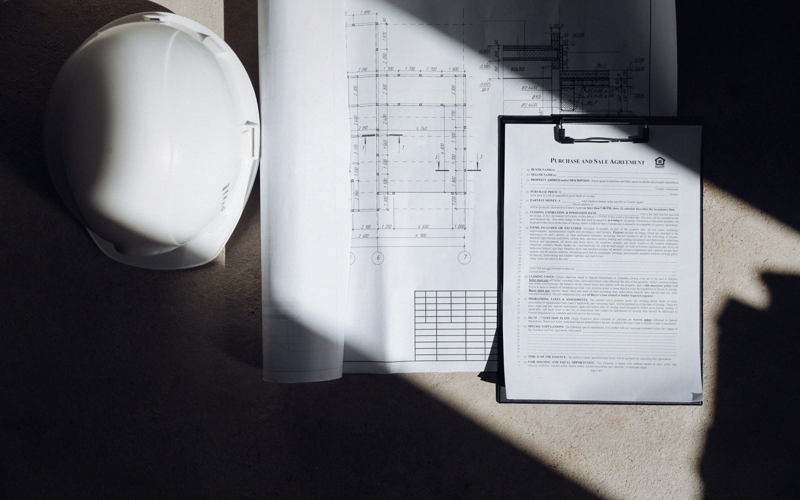 A hard hat, building blueprints, and a purchase and sale agreement on a clipboard