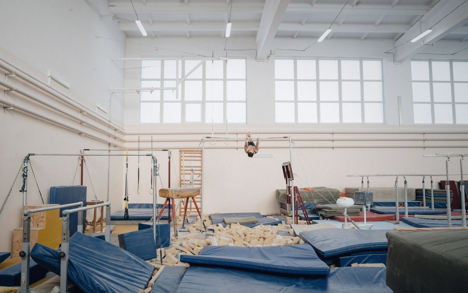 A gymnast performs upside down on a horizontal bar above blue crash mats