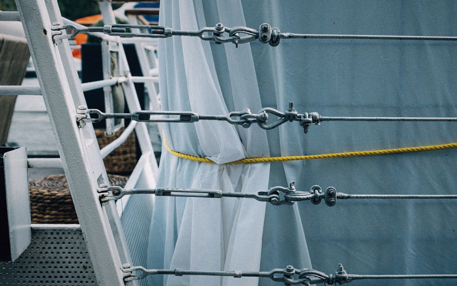 Turnbuckles tension a boat's wire rigging beside a white canvas curtain and yellow rope